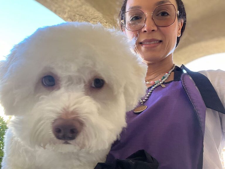 IMG_7275 A woman smiling holds a fluffy white dog. She wears glasses and a purple top, with a beaded necklace.