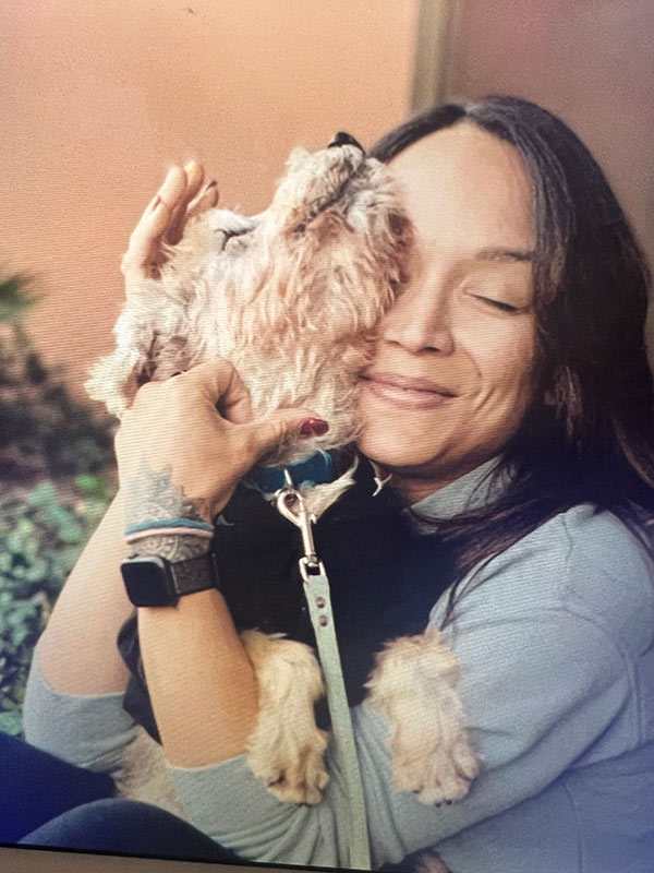 IMG_9690 A woman with long dark hair and a blue sweater joyfully hugs a small dog.